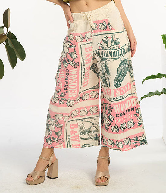 Woman wearing a magnolia horse patterned pants standing in a room with plants.