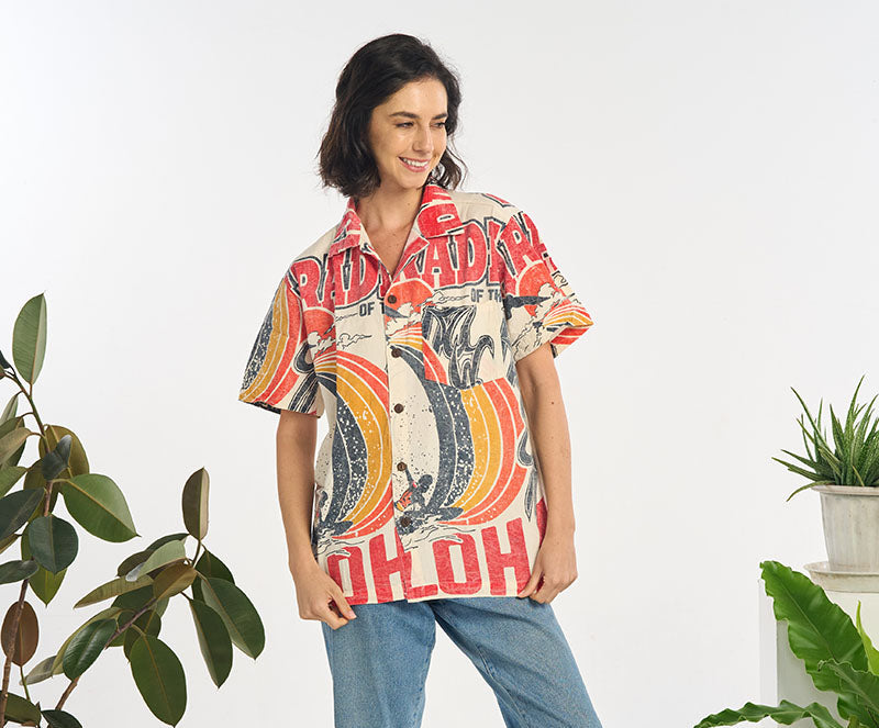 Woman wearing a colorful Aloha shirt and jeans standing in a room with plants.