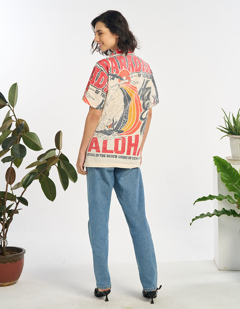 Person wearing a colorful  Aloha shirt with text and graphics, standing in a room with plants.